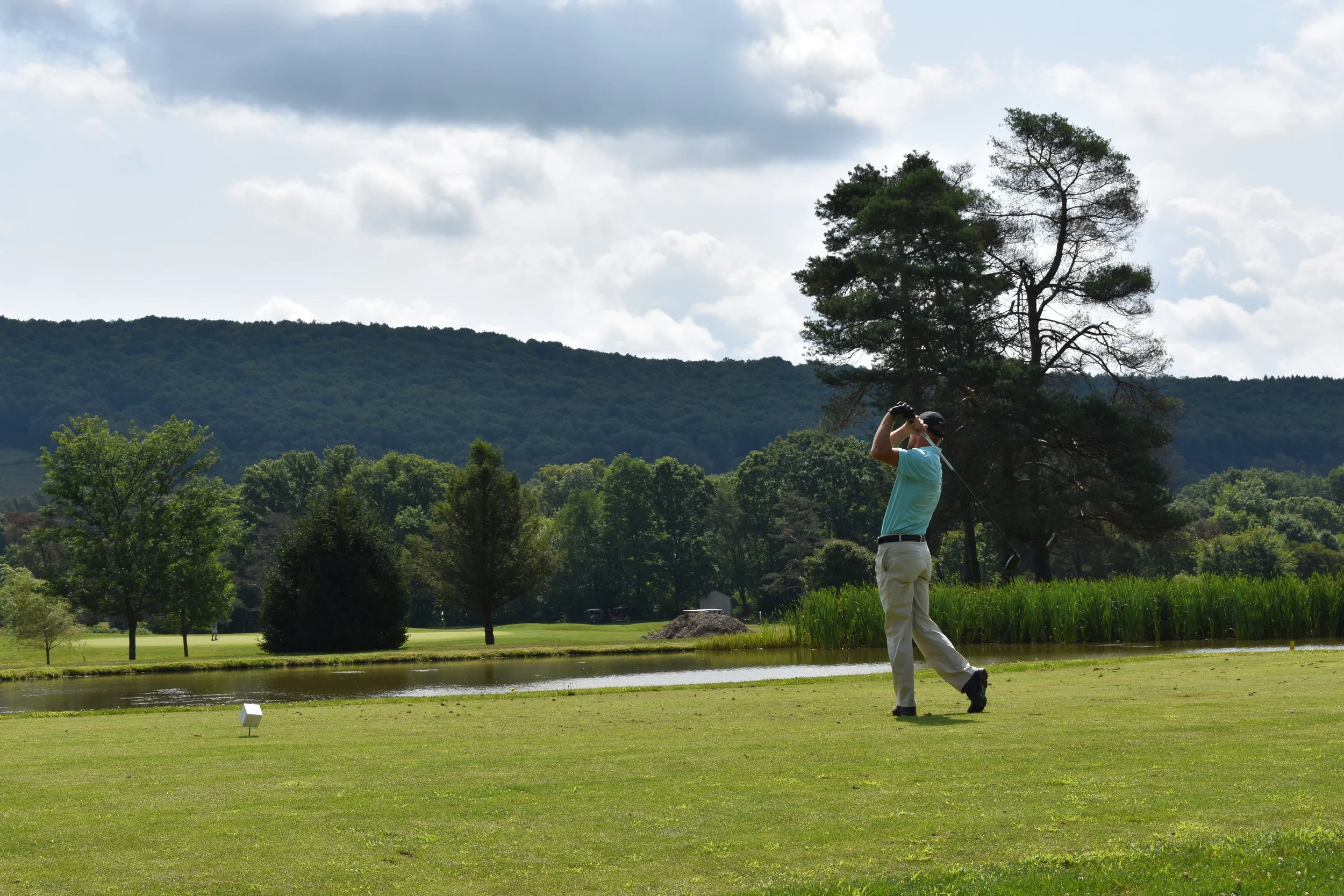 Golfer mid-swing on the tee box with pond in foreground and Chenango Valley mountain ridge under dramatic sky